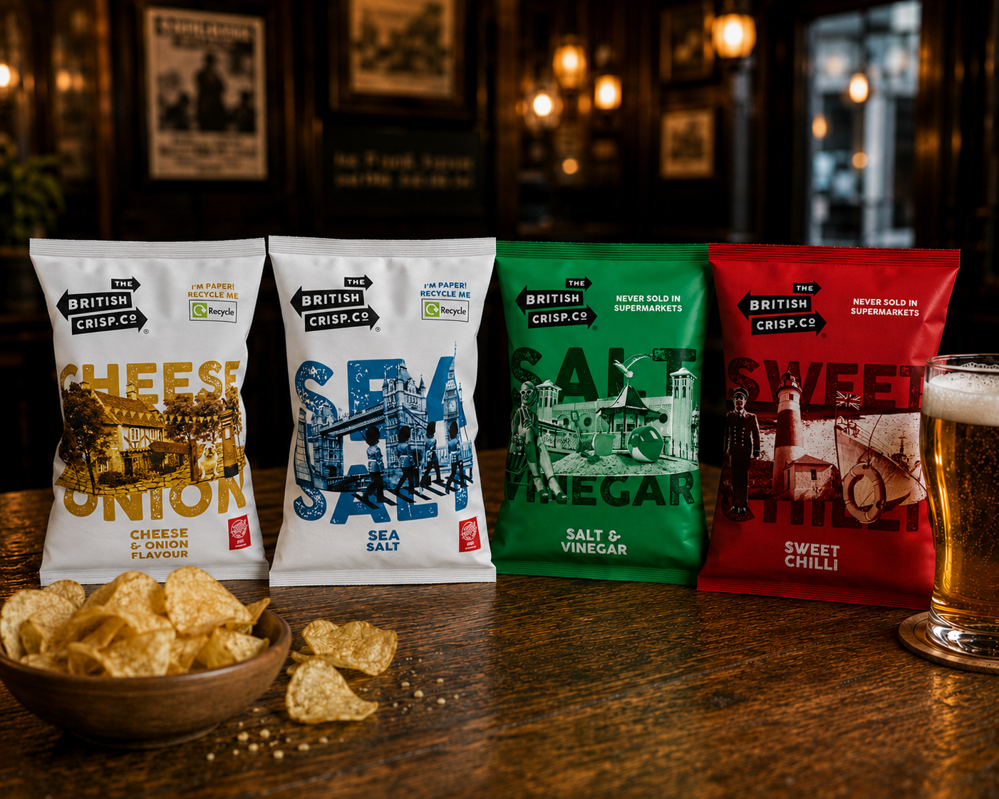 The British Crisp Co. range - two paper packets (Cheese & Onion, Sea Salt) alongside two classic packets (Salt & Vinegar, Sweet Chilli) on a pub bartop with a glass of ale and a bowl of crisps
